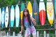 A young woman standing in front of a fence with surfboards.